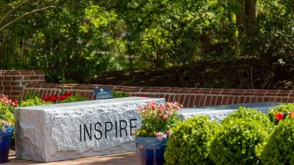 Stone bench engraved with the word “INSPIRE” surrounded by flowers and greenery on campus.