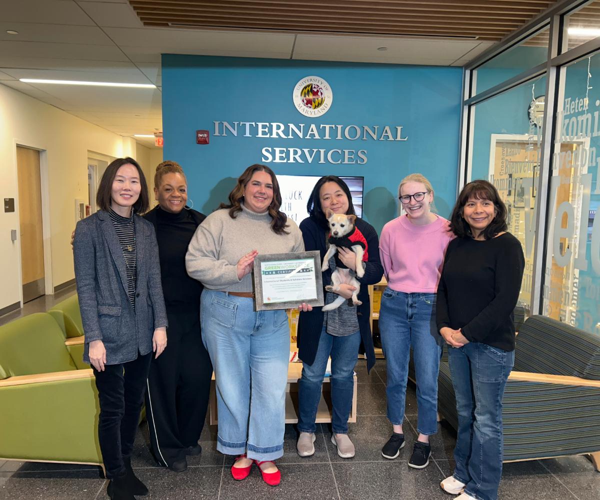 6 staff members, one holding a dog, stand posing with a framed certificate in from of the UMD logo for the Office of International Services