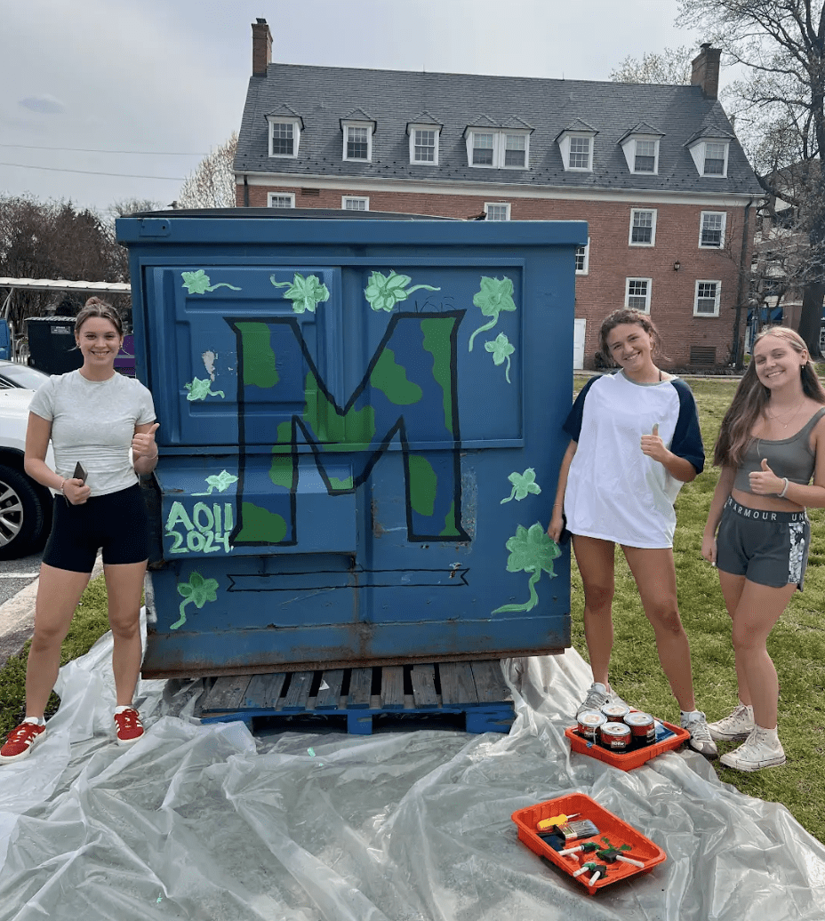 Students stand in front of dumpster painted with an Earth-colored M.