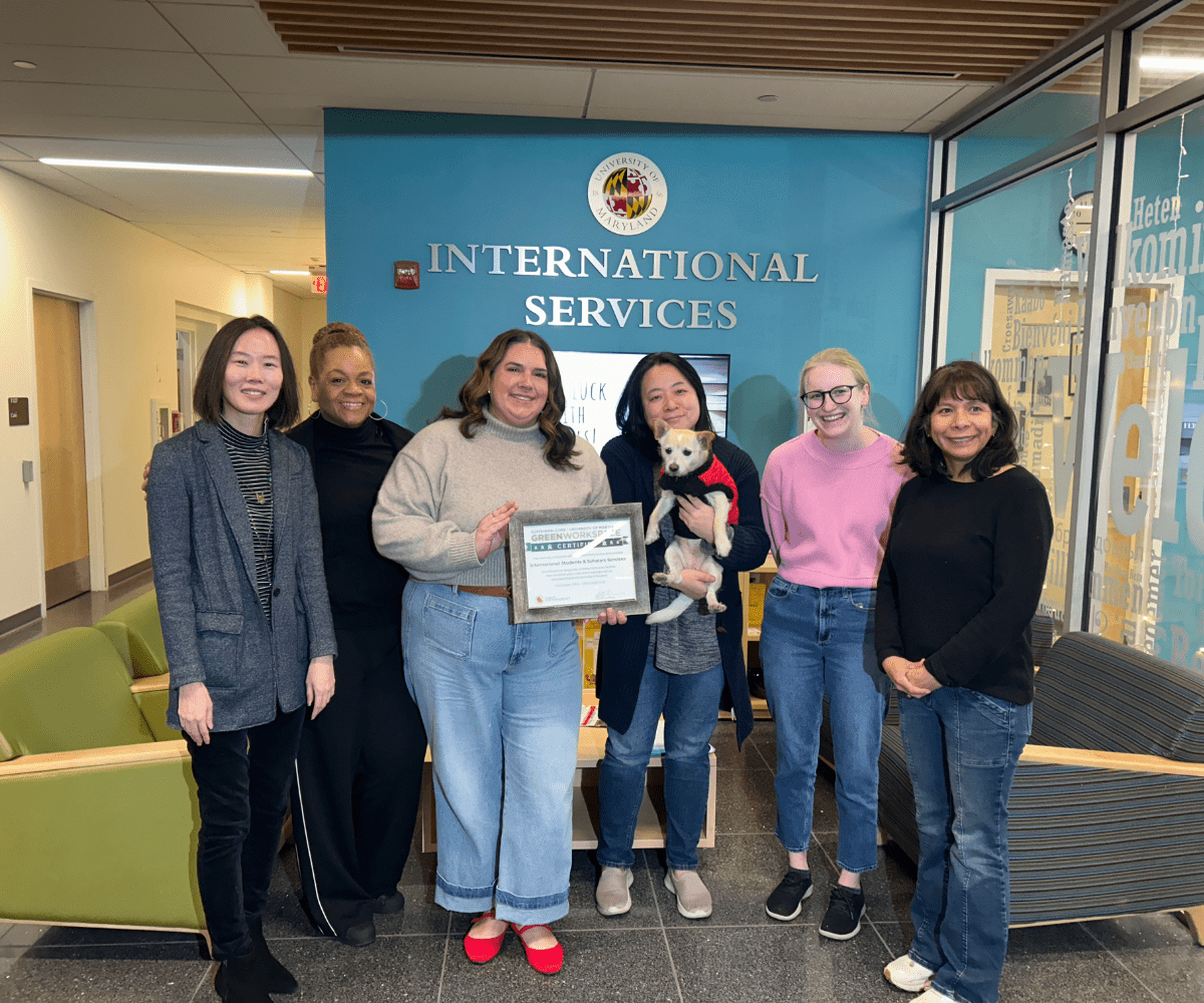 6 staff members, one holding a dog, stand posing with a framed certificate in from of the UMD logo for the Office of International Services