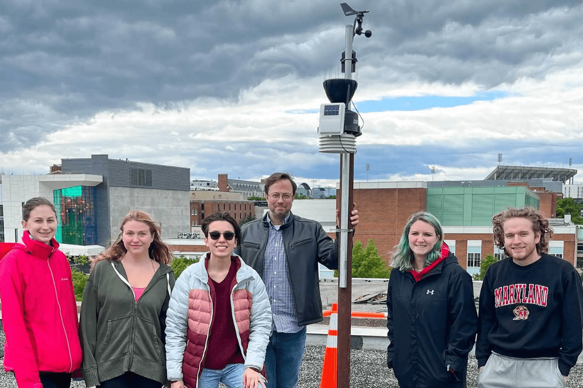 Five students and one faculty stand on the top of a campus building in front of weather equipment