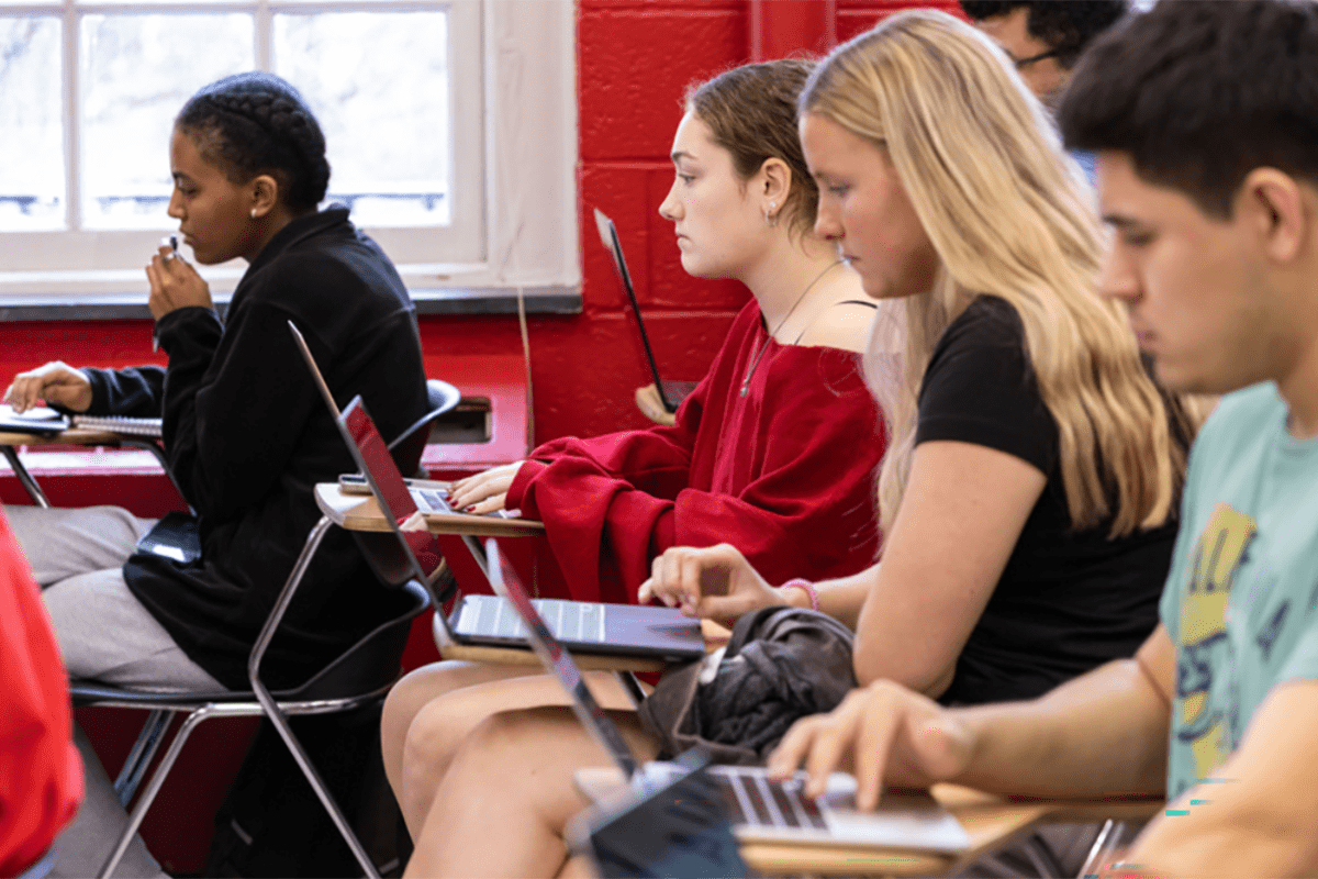 Four students sit in a line of desks in a classroom. All are actively looking at their laptop screens. Red wall in background
