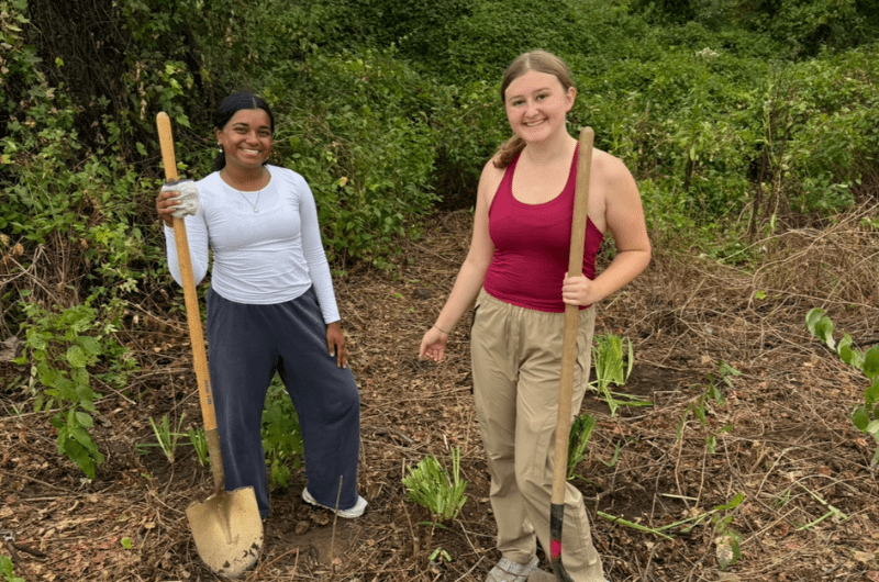 Two students working in an area with dirt and vegetation pause to take a photo holding their shovels