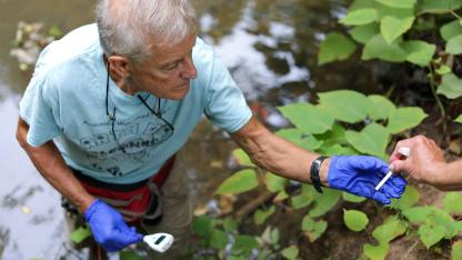 Person wearing gloves collecting a water sample from a stream while another person hands over equipment.