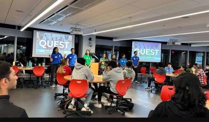 Students gathered at red chairs and tables listening to a presentation in a collaborative learning space.